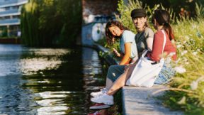 Drei Studierende sitzen am Ufer eines Flusses in einer Stadt und unterhalten sich.