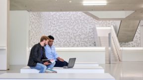 Two students are sitting in front of a laptop in the foyer of the university.